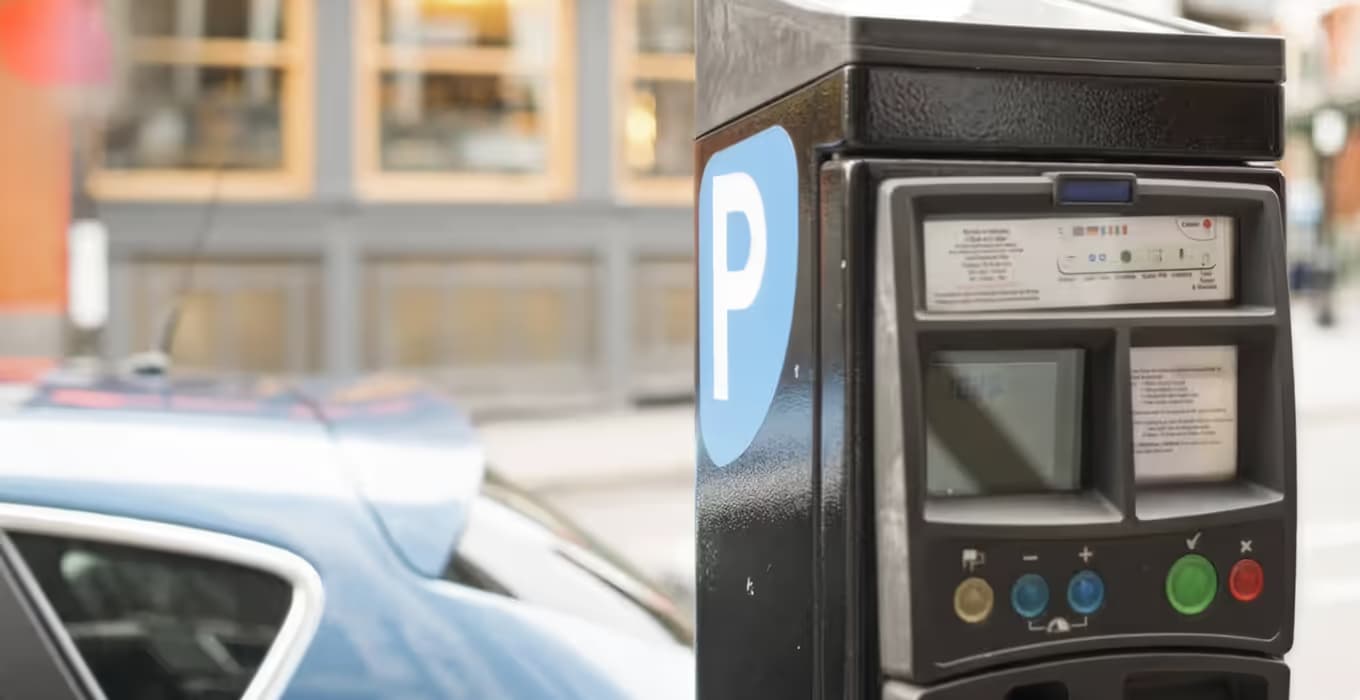 Schwarzer Parkscheinautomat mit blauem P-Schild, dahinter unscharf ein geparktes Auto